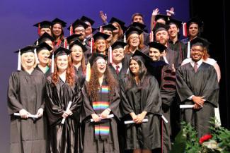 Four rows of smiling students in cap and gown, standing on the Fine Arts stage