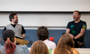 Two men sitting in front of an audience in a classroom. 
