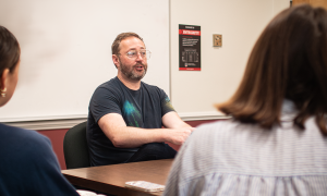A man talks to two students in the foreground. 